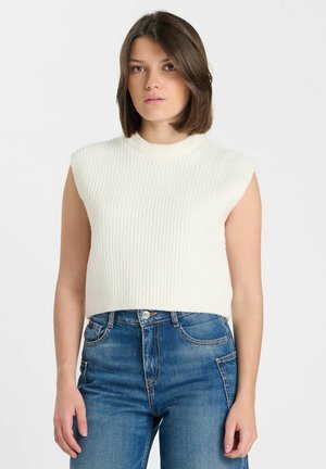 Giovane donna con capelli castani a lunghezza spalle, che indossa un top bianco lavorato a maglia senza maniche e jeans blu a vita alta, in piedi davanti a uno sfondo semplice.