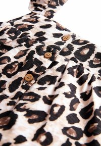 Leopardprint jurk met beige stof en zwarte en bruine vlekken, knoopdetails en verstelbare banden, die een zachte textuur laat zien.