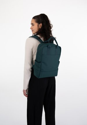 Sac à dos teal avec des bretelles rembourrées, deux poches latérales et des poignées supérieures. Texture lisse, forme rectangulaire et design minimaliste.