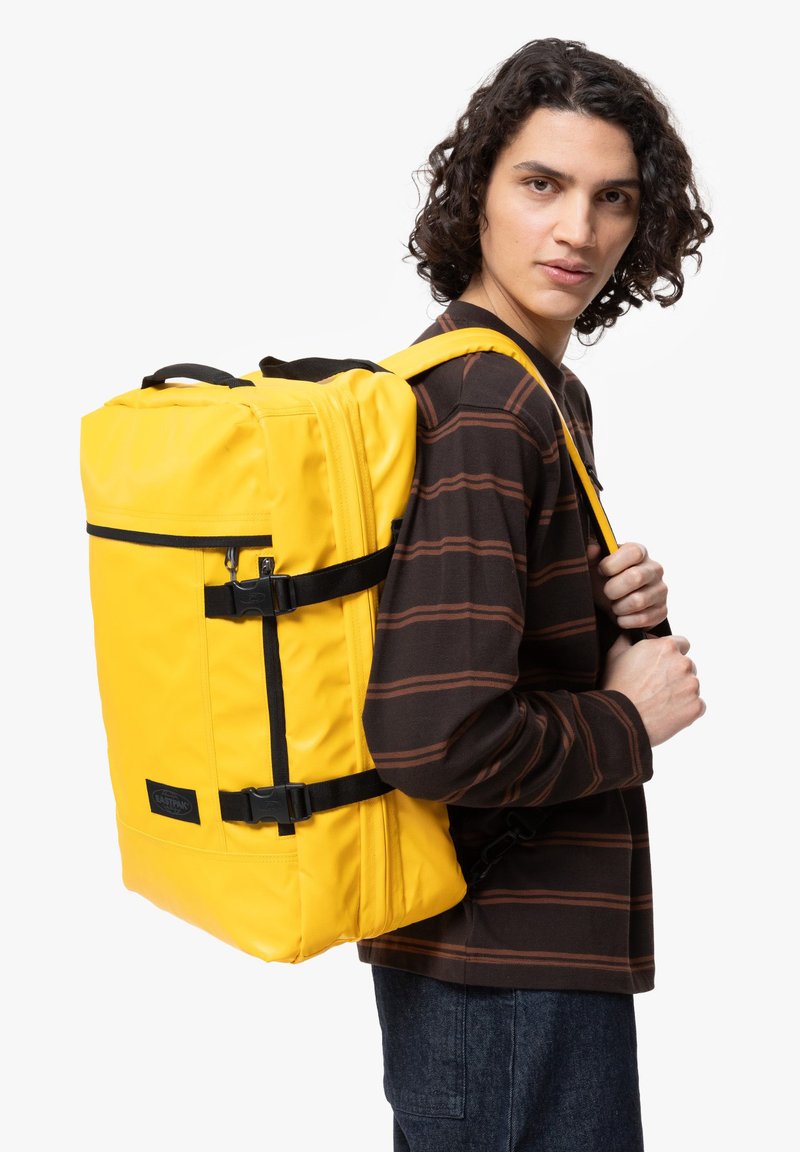 Gul ryggsäck i hållbart material, med justerbara remmar, ett handtag på toppen, sidspännen och en dragkedjeförsedd framficka.