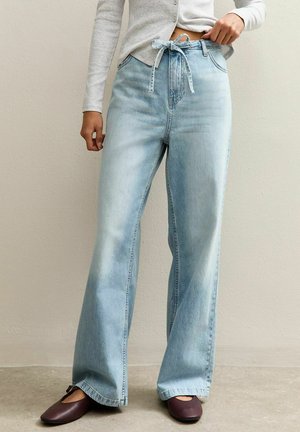 Persoon die lichtblauwe jeans met wijde pijpen en een ceintuur draagt, een grijs lang mouwen top, en donkerbruine platte schoenen op een lichte vloer.