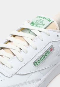 Baskets blanches Reebok avec logo vert et drapeau Union Jack sur le côté, lacets blancs et motif de feuille verte sur l'étiquette de la languette.