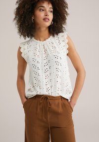 Wit mouwloze blouse met eyelet-patronen, gegolfde randen, textuurstof, gecombineerd met bruine broek met trekkoord.