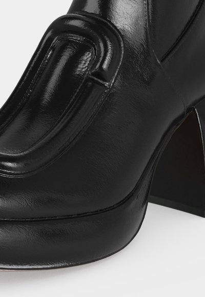 Gros plan d'une chaussure à talon haut en cuir noir brillant avec un talon épais et un panneau décoratif surélevé cousu sur le dessus avant.