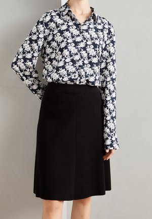 Kobieta ubrana w czarną spódnicę za kolano i granatową koszulę zapinaną na guziki z białym kwiatowym wzorem, stojąca na tle jednolitej szarej ściany.