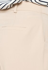 Pantaloni in tessuto beige con una texture liscia e una tasca posteriore con una discreta cucitura. Camicia a righe orizzontali parzialmente visibile sopra.