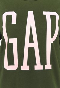 Oliwkowy t-shirt z dużymi, wyraźnymi różowymi literami napisanymi "GAP". Wykonany z miękkiego materiału, z klasycznym okrągłym dekoltem i krótkimi rękawami.
