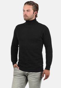 Schwarzer Rollkragensweater mit strukturiertem Gewebe, gerippten Bündchen und Saum, kombiniert mit hellgrauen Jeans. Das Design weist eine figurbetonte Silhouette auf.