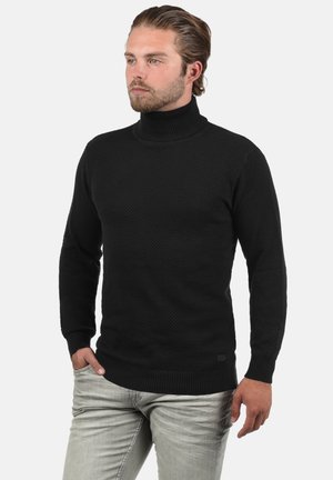 Suéter negro de cuello alto con un tejido texturizado, puños y dobladillo acanalados, combinado con jeans de color gris claro. El diseño presenta una silueta ajustada.
