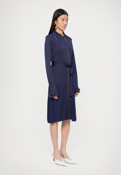 Φόρεμα σε ναυτικό μπλε με πορτοκαλί γιακά και περιθώριο, διαθέτει μακριά μανίκια, ζώνη στη μέση και κλείσιμο με κουμπιά. Παπούτσια σε παστέλ χρώματα συμπληρώνουν την εμφάνιση.