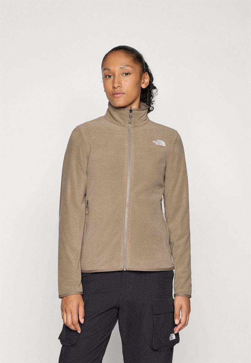 Femme portant une veste en polaire taupe à zip et un pantalon cargo noir avec des logos sur les deux, se tenant devant un fond clair.