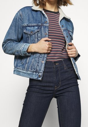 Person iført en blå denimjakke med sherpa-krave over en stribet skjorte og mørke højtaljede jeans, justerer jakken med begge hænder.