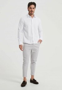 Chemise blanche à manches longues avec col boutonné, associée à un pantalon gris clair et des mocassins en daim marron foncé. Tissu lisse, coupe ajustée.