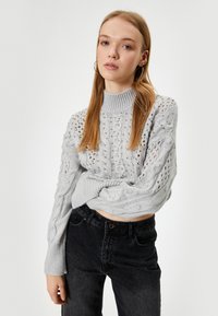 Pull en tricot gris avec un col haut, présentant des motifs en torsades et de la dentelle. Associé à un jean foncé, mettant en avant une coupe décontractée.