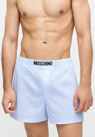 Άνδρας με κορμό που φοράει ανοιχτό μπλε και άσπρο ριγέ μποξεράκια Moschino με ελαστική μέση και λογότυπο μπροστά στο κέντρο.