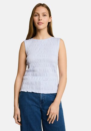 Młoda kobieta ubrana w bezrękawnik w jasnoniebieskim, teksturowanym materiale oraz niebieskie dżinsy, stojąca na tle jednolitego, białego tła.