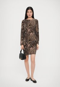 Φόρεμα μακρυμάνικο με καφέ paisley σχέδιο σε υφή ύφασμα. Το μοντέλο κρατά μια μικρή μαύρη τσάντα χεριού και φορά μαύρες επίπεδες παπουτσάκια.