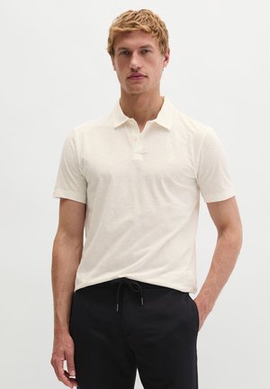 Młody mężczyzna stoi, ubrany w kremową koszulkę polo z krótkim rękawem i czarne spodnie z troczkami na jednolitym jasnym tle.