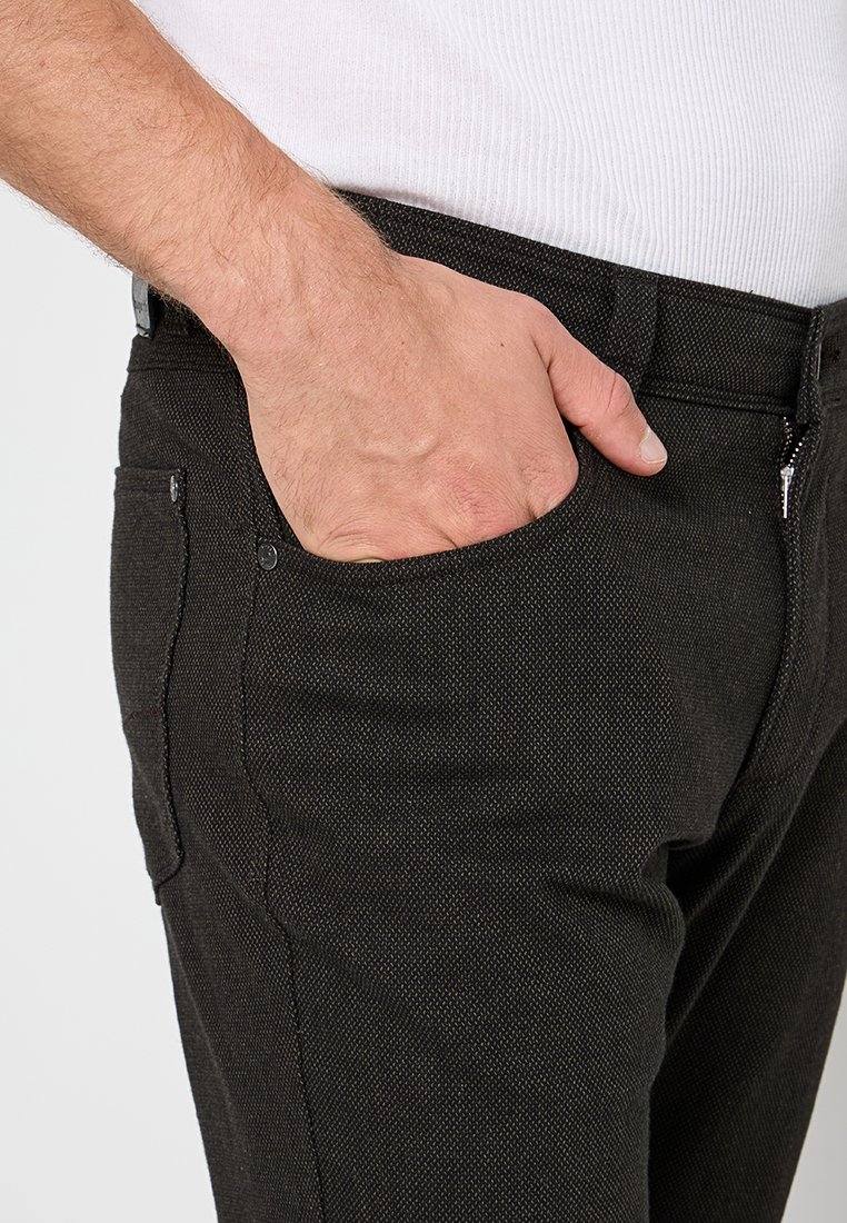 Rechte Hand in der Vordertasche von dunkel strukturierten Hosen, die mit einem weißen, gerippten Shirt getragen werden.