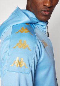 Haut à capuche bleu clair avec des graphiques dorés sur les épaules dans un motif répété, fabriqué à partir d'un tissu lisse et texturé avec une fermeture éclair.