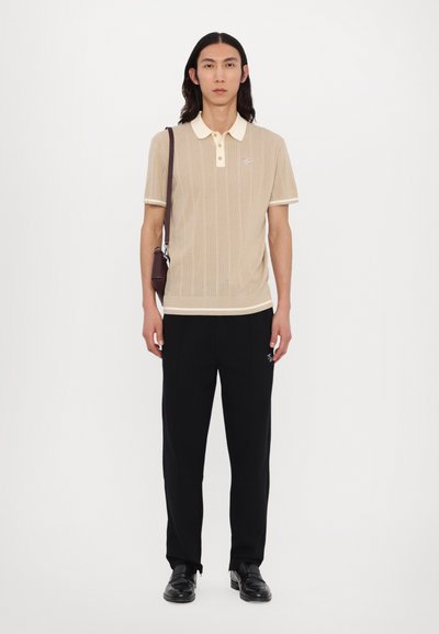 Beige polo tričko s krátkým rukávem a texturovaným vzorem, smetanový límeček a manžety, doplněné o černé kalhoty a černé boty. Tmavá kabelka přes rameno.