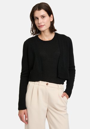 Zwarte cropped cardigan van zachte stof, met lange mouwen en een open voorkant, gecombineerd met lichtbeige geplooide broeken.