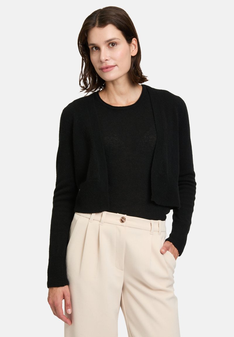 Gilet noir court en matériau doux, avec des manches longues et une ouverture à l'avant, associé à un pantalon plissé beige clair.