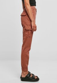 Cargo-byxor i rostfärg, tillverkade av bomull. Har elastiska muddar, sidofickor och en åtsittande design. Kombineras med svarta flip-flops.
