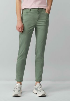 Person trägt eng anliegende grüne Hose, hellrosa Hemd und weiße-graue Sneakers, steht mit einer Hand in der Tasche.