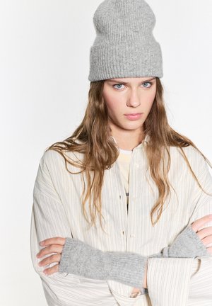 Grijze geribbelde gebreide beanie en polswarmers, gecombineerd met een beige gestreepte oversized shirt. Model heeft lang haar en heeft haar armen voor zich gekruist.