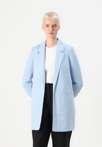 Απαλή μπλε σακάκι κομμένο με εσοχή, κούμπωμα με έναν κουμπί και δύο πλευρικές τσέπες, φορεμένο πάνω από λευκό τοπ και μαύρο παντελόνι.