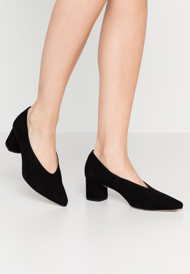 Escarpins en daim noir avec un bout pointu, un talon carré moyen, et un design à découpe en forme de V sur les côtés. Texture lisse et style minimaliste.