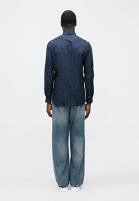 Námorníckomodrá košeľa s dlhými rukávmi a geometrickým vzorom, v kombinácii s bledomodrými džínsami s širokými nohavicami a bielymi teniskami. Minimalistický dizajn, hladká textúra.