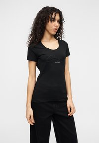 Μαύρο T-shirt με στρογγυλή λαιμόκοψη και κοντά μανίκια, με ανάγλυφο λογότυπο "Guess" και "est. 1981" κεντημένο με αντίθετα χρώματα.