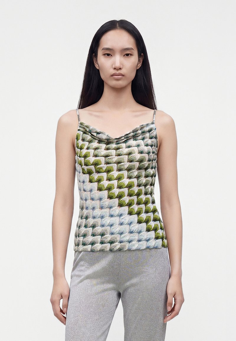 Femme aux longs cheveux noirs portant un haut sans manches multicolore texturé et un pantalon gris clair, se tenant devant un fond uni.