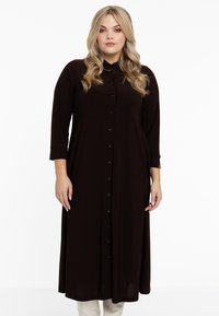 Robe longue marron foncé en tissu doux, avec boutons sur le devant, col et manches trois-quarts. Texture lisse et design simple.