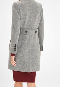 Manteau en laine gris avec une coupe ajustée, comportant un détail double bouton au dos et des manches boutonnées. Le manteau est associé à une robe bordeaux à côtes.