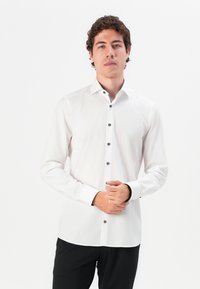 Camisa blanca de manga larga con botones negros, cuello puntiagudo y acentos negros en contraste en los puños y la tapeta; tejido suave y ligero.