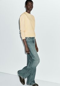 Light yellow sweater, relaxed fit, ribbed cuffs. Faded blue flared jeans, denim texture, with subtle stitching details. Brown shoes.