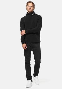 Fekete bolyhos garbó, hajtott gallérral, fekete slim-fit farmernadrággal és fehér tornacipővel párosítva, egy kis hímzett mintával.