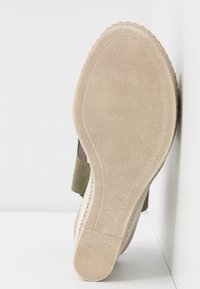Semelle d'une chaussure en daim beige avec un dessous à texture lisse et des coutures visibles, posée contre une surface blanche.