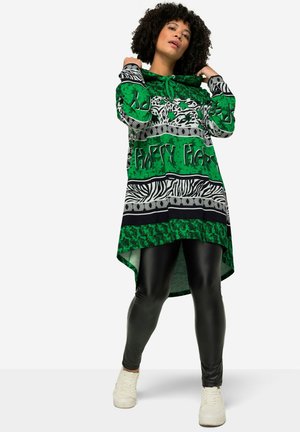 Tunique longue à motifs verts avec des rayures zèbres noires et blanches et du texte, associée à des leggings noirs brillants et des baskets blanches.