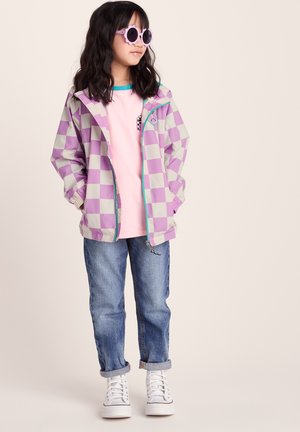 Young girl wearing purple and white checkered jacket, pink shirt, blue jeans, white sneakers, and round purple sunglasses standing against plain background.