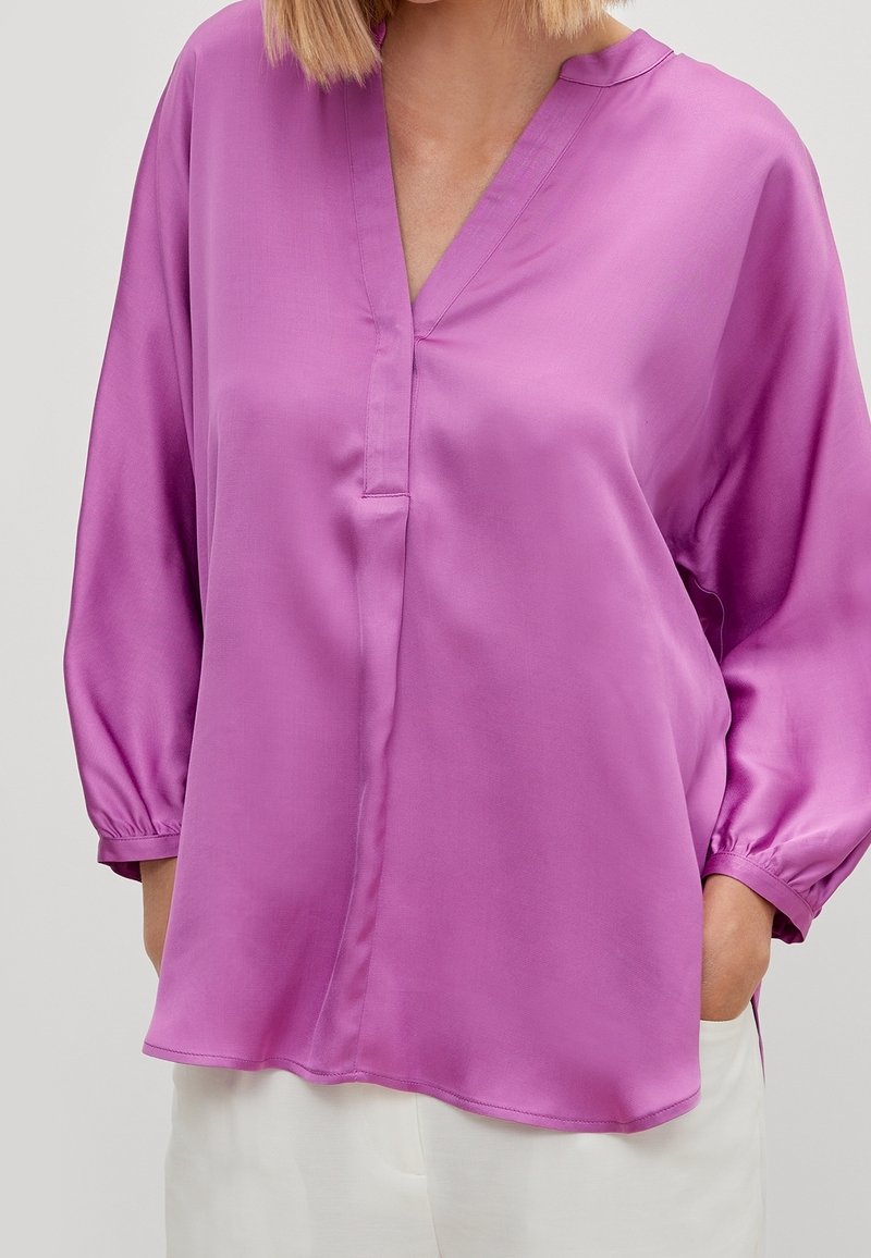 Femme portant un chemisier violet ample à manches longues avec un col en V et un pantalon blanc, mains dans les poches, sur un fond uni.