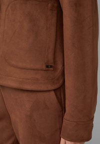 Giacca in suede marrone con una grande tasca e un piccolo logo sul lato sinistro. Il materiale ha una finitura morbida e testurizzata.
