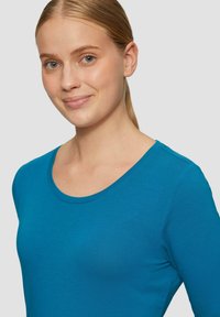 Langarmige teal shirt met een ronde hals en een zachte textuur, getoond op een model met steil haar tegen een neutrale achtergrond.
