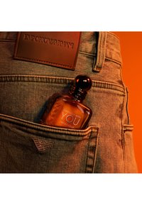 Bottiglia di profumo in vetro marrone etichettata "Stronger With You Intensely" nella tasca frontale dei jeans con loghi in pelle e metallo Emporio Armani.
