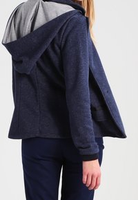 Marinblå hoodie med gråfoder, strukturerad tyg, avslappnad passform och ribbade ärmslut. Har en stor huva och något öppen fram.