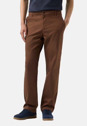 Man draagt recht vallende bruine broek met handen in de zakken en marineblauwe sneakers op een witte achtergrond.