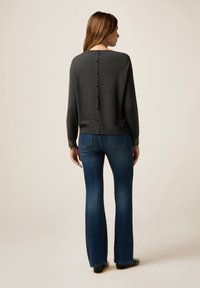 Grå stickad tröja med rund hals, knapprad på baksidan och kontrasterande texturerade ärmar, kombinerad med utsvängda blå jeans.